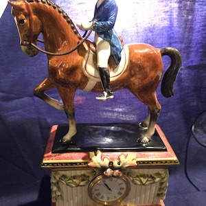 May include: A ceramic mantel clock featuring a brown horse with a rider in a blue jacket and top hat. The base is rectangular with a white face, black Roman numerals, and decorative leaf motifs in shades of red, green, and gold around the clock.