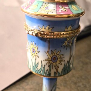 Pode incluir: Uma caixa de bugigangas decorativa de porcelana pintada à mão em forma de caixa de correio. A caixa apresenta um topo vermelho, uma tampa azul e branca e um corpo cilíndrico adornado com girassóis e abelhas. A base é rosa com desenhos de abelhas e folhas.