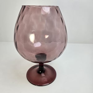 Puede incluir: Un vaso de brandy vintage de cristal color amatista. El cuenco tiene una superficie texturizada y se asienta sobre un tallo corto y una base circular. El cristal es translúcido.