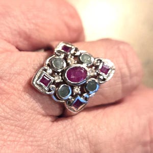 May include: A silver-toned ring featuring a central oval-shaped deep pink gemstone surrounded by four round grey-green stones and four smaller square-cut deep pink stones, accented with tiny clear stones set in a decorative metal frame.