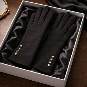 May include: A pair of black gloves with gold-colored button details, presented in a white box lined with black satin. The gloves are made of a soft material and are a classic accessory.