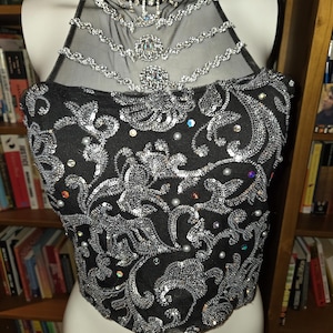Puede incluir: Top halter negro con adornos de lentejuelas y cuentas plateadas. El top presenta un diseño floral y un escote transparente con detalles de pedrería. El corpiño está adornado con lentejuelas y pequeñas perlas, creando un look glamuroso.