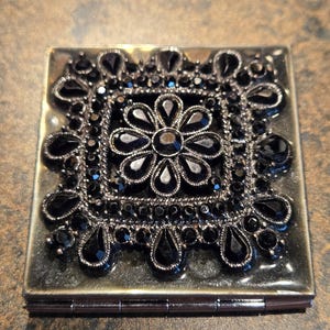 May include: A square, silver-toned compact mirror with a black floral design. The design features a central flower and surrounding black teardrop-shaped embellishments. The compact is closed and has a hinged closure.