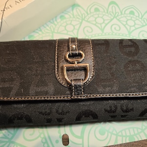Puede incluir: Cartera de mano negra con un patrón repetido y cierre de solapa de cuero. La cartera presenta un cierre de metal dorado y un borde cosido. La marca "a Etienne Aigner" es visible en la caja del fondo.