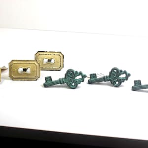 May include: Three rectangular, cream-colored metal keyhole plates with distressed edges and three teal-colored metal key-shaped drawer pulls featuring ornate, filigree-style handles and a textured, weathered finish on the surface.