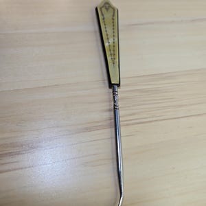 May include: An antique crochet hook with a decorative handle. The handle is cream-colored with a black border and a detailed design. The hook is made of silver-colored metal and has a curved end.