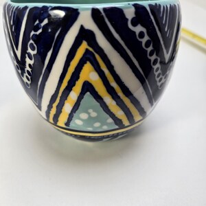 May include: A ceramic bowl with a geometric pattern in navy blue, white, and yellow. The interior is a light teal color. The bowl has a rounded square shape.