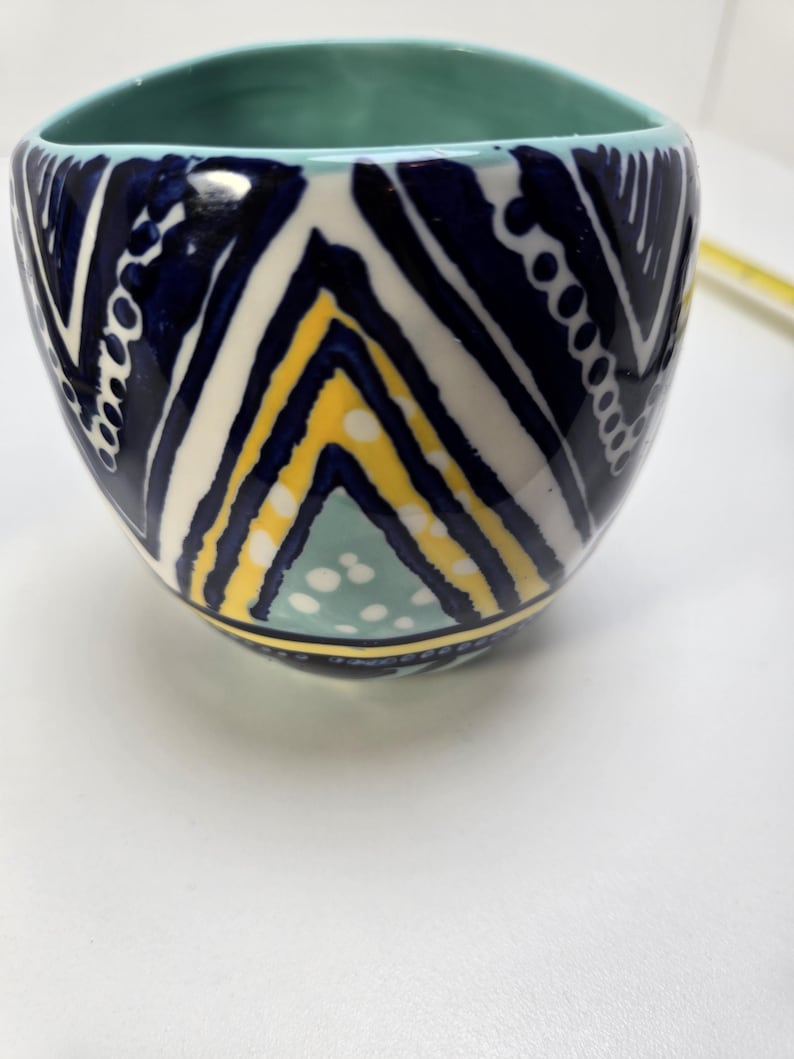 May include: A ceramic bowl with a geometric pattern in navy blue, white, and yellow. The interior is a light teal color. The bowl has a rounded square shape.