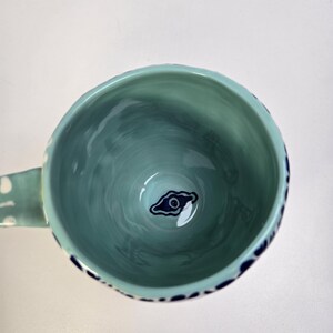 May include: A teal ceramic mug with a handle, featuring a dark blue eye design at the bottom. The interior has a marbled effect, and the rim is decorated with dark blue dots. The handle has white polka dots.
