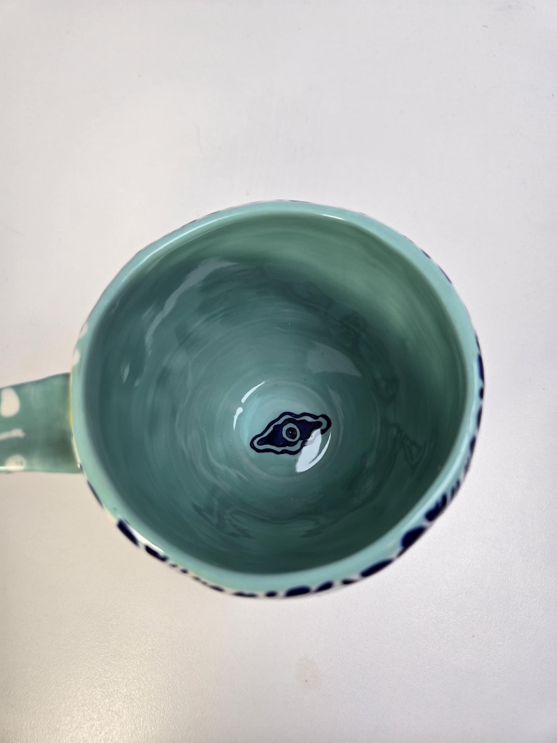 May include: A teal ceramic mug with a handle, featuring a dark blue eye design at the bottom. The interior has a marbled effect, and the rim is decorated with dark blue dots. The handle has white polka dots.
