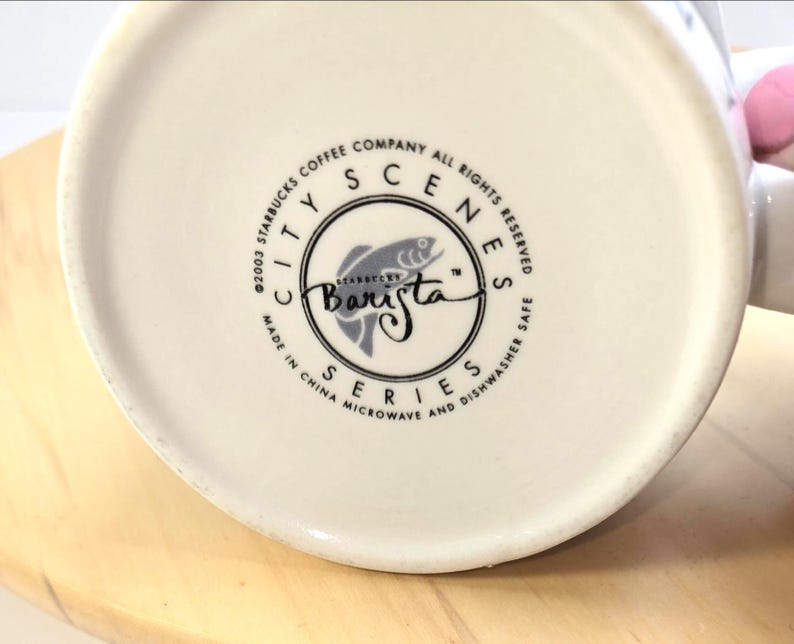May include: Off-white ceramic mug with a circular logo featuring a fish illustration and the word "Barista." The logo is surrounded by text that reads "City Scenes Series" and "Made in China." The mug is microwave and dishwasher safe.
