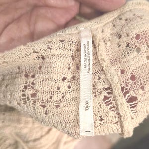 Op de afbeelding: Close-up van een cr&egrave;mekleurig, opengebreid kledingstuk. De stof heeft een delicaat, kantachtig patroon met kleine gaatjes. Een wit label leest "MADE IN CHINA FABRIQU&Eacute; EN CHINE" en "S P".
