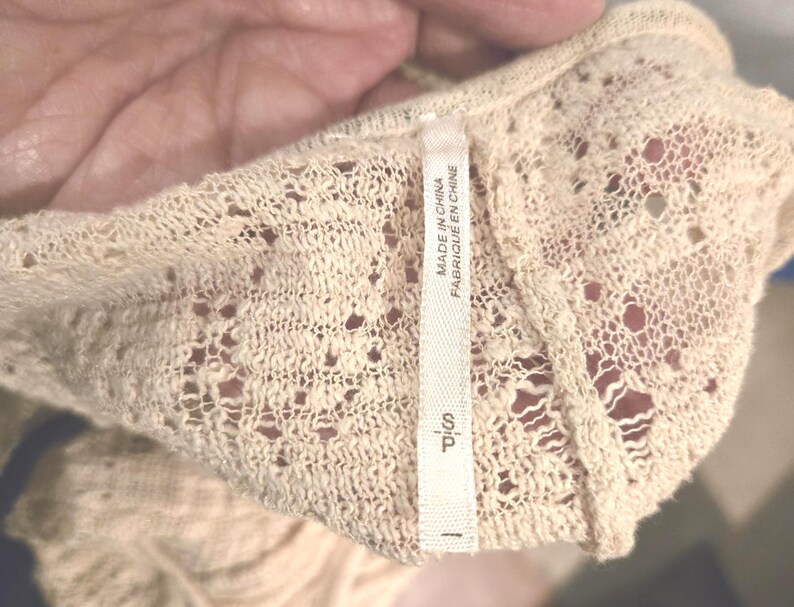 Op de afbeelding: Close-up van een cr&egrave;mekleurig, opengebreid kledingstuk. De stof heeft een delicaat, kantachtig patroon met kleine gaatjes. Een wit label leest "MADE IN CHINA FABRIQU&Eacute; EN CHINE" en "S P".