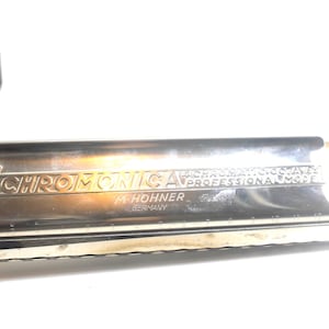 Könnte beinhalten: Eine silberfarbene M. Hohner Chromonica Mundharmonika mit 64 chromatischen Tönen, Profimodell, hergestellt in Deutschland. Das Metallgehäuse trägt die Gravur The 64 Chromonica 4 Chromatic Octaves Professional Model M. Hohner Germany.