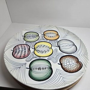 May include: A white ceramic plate with a hand-painted design featuring six stylized fruit halves in various colors. The plate has a black and white geometric pattern around the edge and a wooden base.