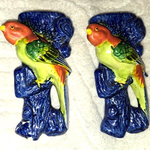 May include: A pair of ceramic wall plaques featuring colorful parrots with orange heads, green wings, and yellow bodies perched on textured, dark blue tree stumps. The birds have small yellow beaks and are depicted in a side profile view.