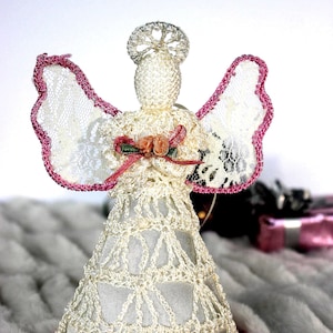 May include: A white crocheted angel ornament with pink lace wings and a pink satin bow with a floral accent. The angel is sitting on a white fluffy surface.