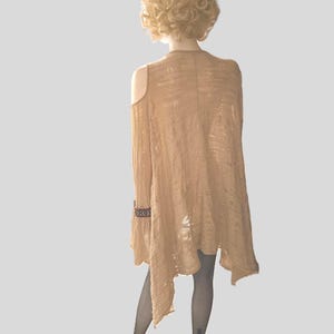Op de afbeelding: Een lichtbruin vest met open schouders en een asymmetrische zoom. De lange mouw heeft een decoratieve manchet. Het kledingstuk wordt getoond op een mannequin met een blonde pruik en zwarte visnetkousen. Kleding.