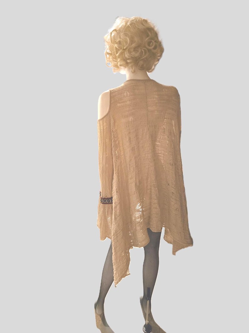 Op de afbeelding: Een lichtbruin vest met open schouders en een asymmetrische zoom. De lange mouw heeft een decoratieve manchet. Het kledingstuk wordt getoond op een mannequin met een blonde pruik en zwarte visnetkousen. Kleding.