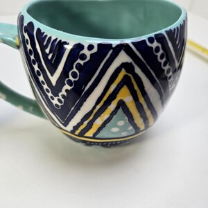 May include: A ceramic mug with a teal interior and handle. The exterior features a navy blue, white, and yellow geometric pattern. The mug has a wide, rounded shape and is set against a white background.