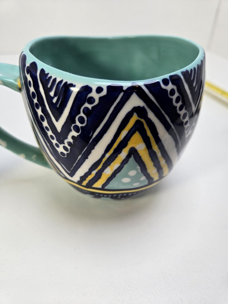 May include: A ceramic mug with a teal interior and handle. The exterior features a navy blue, white, and yellow geometric pattern. The mug has a wide, rounded shape and is set against a white background.