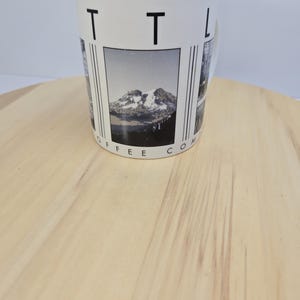May include: White ceramic coffee mug with a mountain landscape design. The mug features the text "TTL COFFEE CO" in black. The mountain image is in black and white. The mug is on a light brown wooden surface.