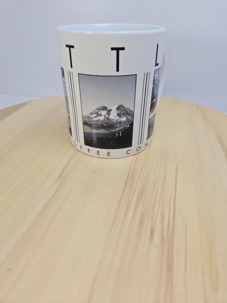 May include: White ceramic coffee mug with a mountain landscape design. The mug features the text "TTL COFFEE CO" in black. The mountain image is in black and white. The mug is on a light brown wooden surface.