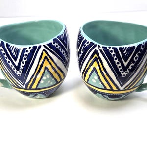 May include: Two ceramic mugs with a teal interior and handles. The exterior features a geometric pattern in navy blue, white, and yellow. The handles are teal with white polka dots. These mugs are ideal for coffee or tea.