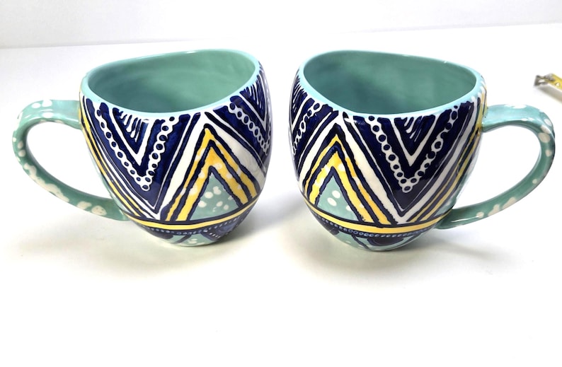 May include: Two ceramic mugs with a teal interior and handles. The exterior features a geometric pattern in navy blue, white, and yellow. The handles are teal with white polka dots. These mugs are ideal for coffee or tea.