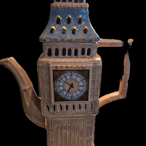 Puede incluir: Una tetera de cerámica con forma de la torre del reloj Big Ben. La estructura es de color beige claro con un techo azul y detalles dorados. Una esfera de reloj cuadrada con números romanos negros está centrada en la parte frontal. Pico y asa curvos.