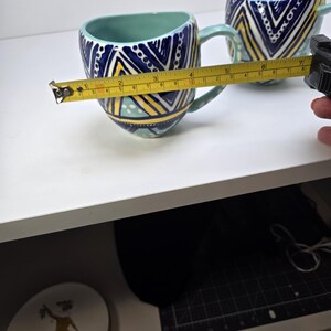 May include: Two ceramic mugs with a teal interior and handle. The exterior features a geometric pattern in navy blue, yellow, and white. The mug is approximately 4 inches wide. The mugs are on a white surface.