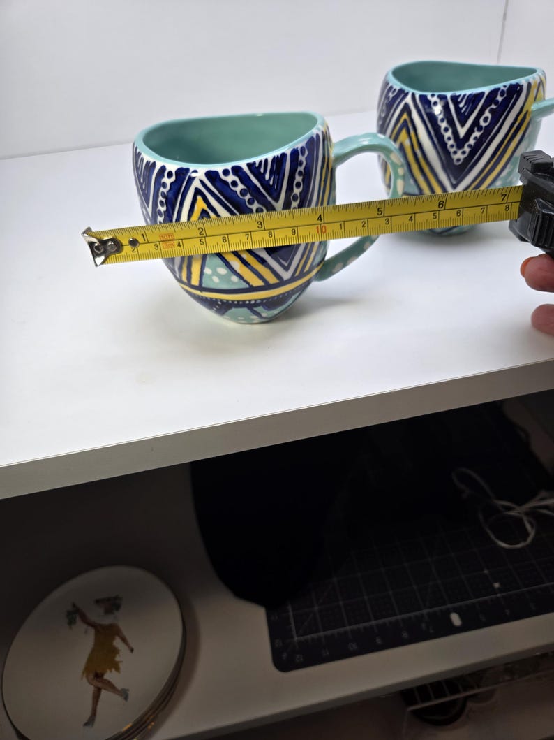 May include: Two ceramic mugs with a teal interior and handle. The exterior features a geometric pattern in navy blue, yellow, and white. The mug is approximately 4 inches wide. The mugs are on a white surface.
