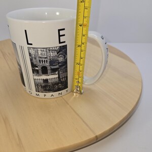 May include: White ceramic mug with a black and white image of a building and the text "LE COMPANY". The mug is approximately 4 inches tall. The mug is on a light brown wooden surface.