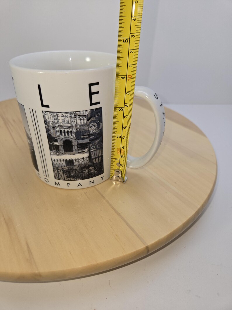 May include: White ceramic mug with a black and white image of a building and the text "LE COMPANY". The mug is approximately 4 inches tall. The mug is on a light brown wooden surface.