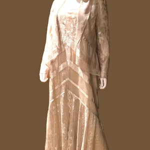 May include: A floor-length beige dress with a matching long-sleeve jacket. The ensemble features a delicate floral lace overlay, a V-neckline on the jacket, and horizontal paneling details on the skirt that create a layered, textured appearance.