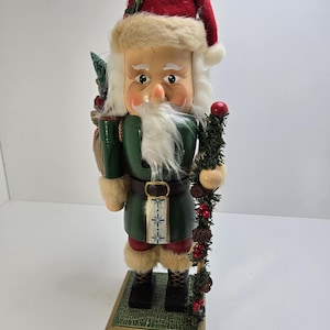 May include: A wooden Christmas nutcracker figurine in the shape of Santa Claus. The figurine wears a red hat, green tunic, and black boots. It holds a staff with pinecones and berries. The base is green and gold with the year "2002" printed on it.