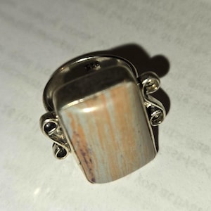 May include: A silver ring featuring a rectangular, vertically striped stone in shades of tan, cream, and grey. The metal setting includes decorative scrollwork on both sides of the stone, and the interior of the band is stamped with the number 925.