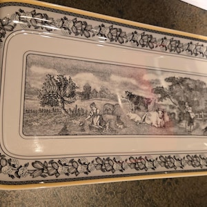 Puede incluir: Una bandeja de servicio rectangular de cerámica con base color crema y borde amarillo. El centro presenta una escena pastoral en blanco y negro con vacas, una persona y un edificio. Un patrón floral rodea la ilustración central.