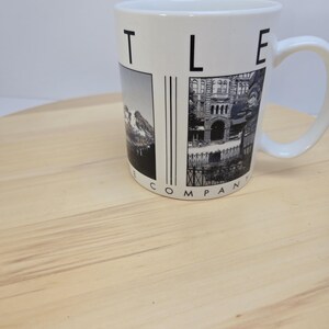 May include: White ceramic mug with a black and white image of a mountain range and a building. The mug has a curved handle and the word "COMPANY" is printed on the side. The mug is sitting on a light brown wooden surface.