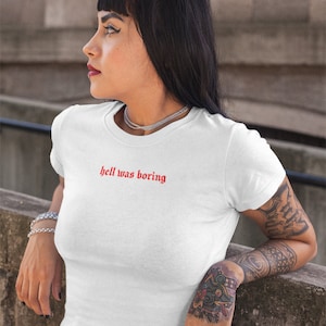 Op de afbeelding: Wit T-shirt met ronde hals en de rode tekst "hell was boring". Het shirt is gemaakt van een zacht materiaal. Het model draagt een zilveren ketting en armband. Het model heeft tatoeages op arm en hand.