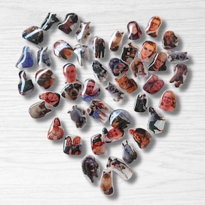 May include: A heart-shaped arrangement of small, glossy magnets on a white wooden surface. Each magnet displays a different photograph, featuring portraits of people and animals. The magnets are of various sizes and shapes, creating a unique and personalized display.