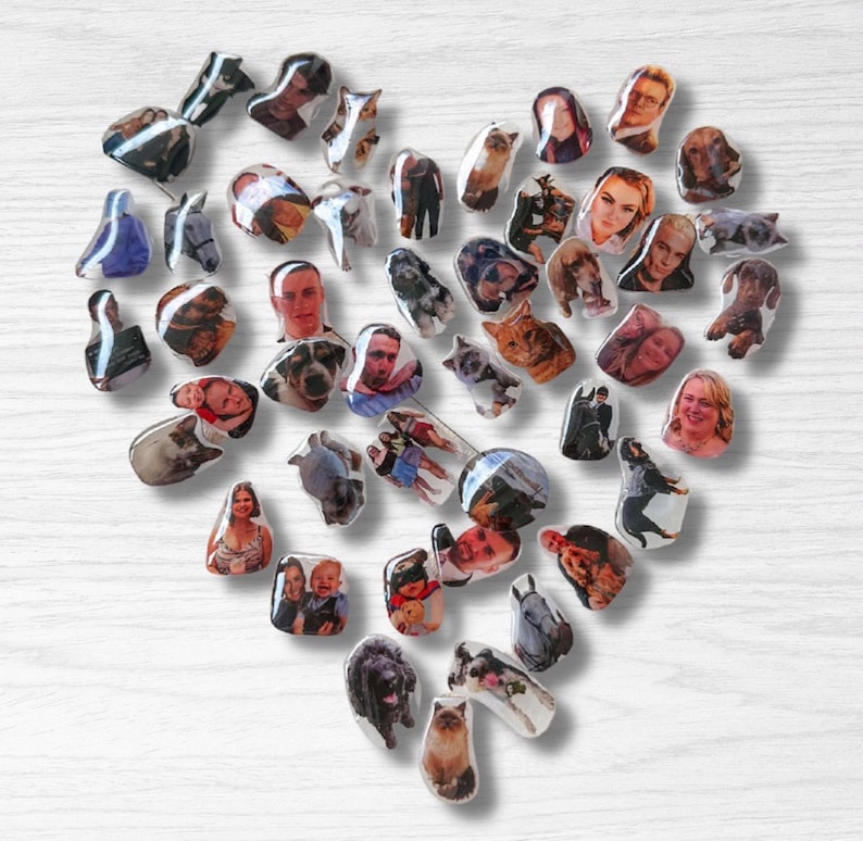 May include: A heart-shaped arrangement of small, glossy magnets on a white wooden surface. Each magnet displays a different photograph, featuring portraits of people and animals. The magnets are of various sizes and shapes, creating a unique and personalized display.