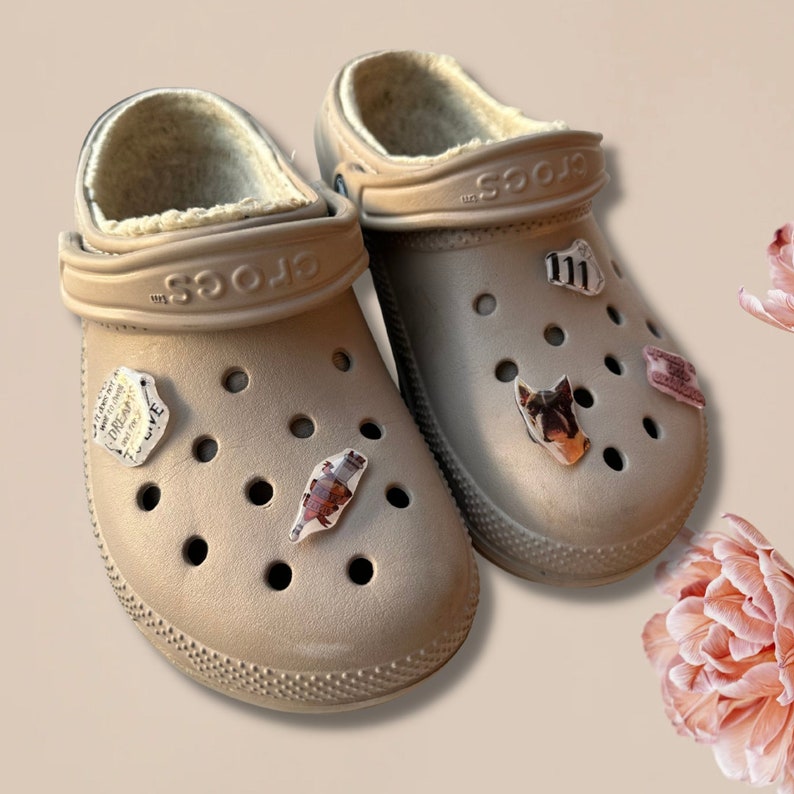 May include: Two tan Crocs clogs with a fluffy inner lining and decorative charms. The charms include a dog's face, text, and other small images. The shoes are on a light tan surface.