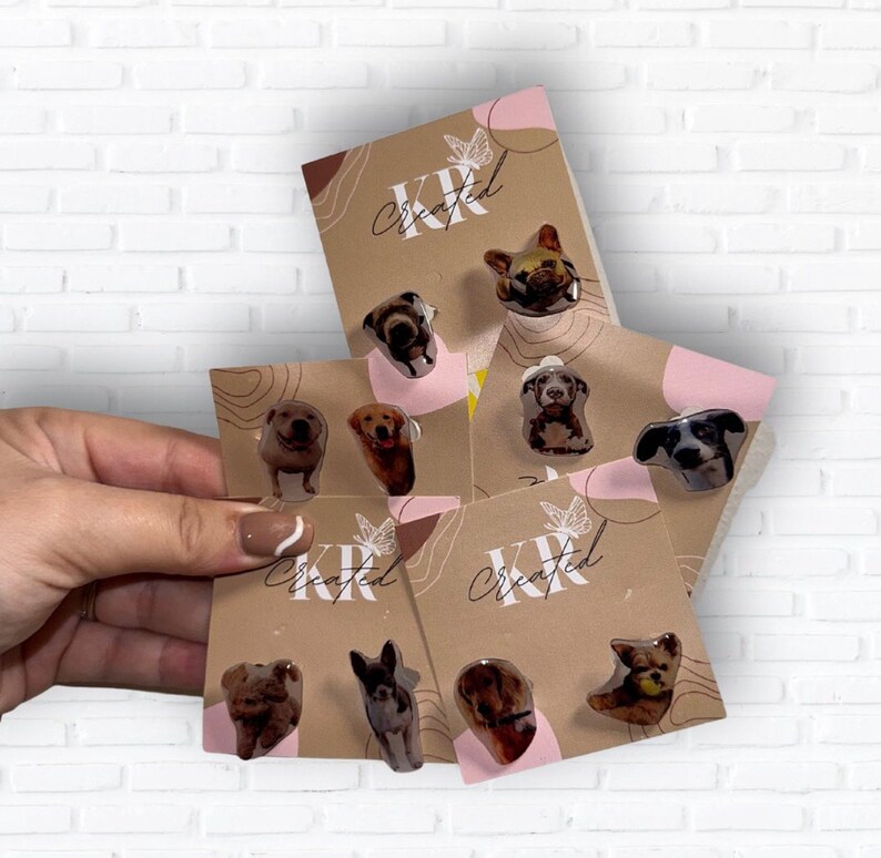 May include: A selection of dog breed stud earrings presented on kraft paper cards. The earrings showcase different dog breeds with a glossy finish. The cards are branded with "KR Created" and feature butterfly and abstract designs.