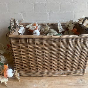 May include: A rectangular wicker basket with woven handles, filled with decorative autumn leaves and small pumpkins. The basket is light brown and sits on a wooden surface, evoking a rustic autumn theme.