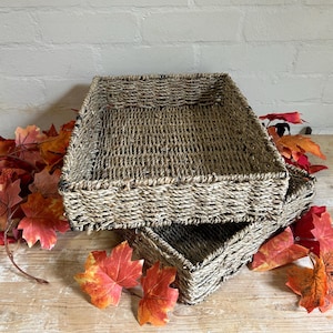 May include: Two rectangular woven baskets made of natural seagrass fibres with a tight, braided texture. The baskets are stacked on top of each other, featuring a light brown and tan colour palette with visible variations in the natural material.