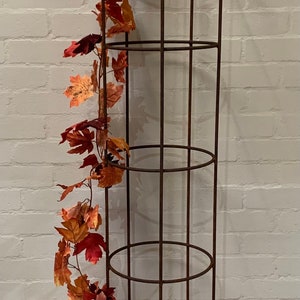 May include: A rusty metal plant stand with four rounded posts and three circular shelves. The stand is decorated with a garland of artificial autumn leaves in shades of orange and red.