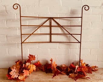 May include: A rusty metal trellis with a curved top and a geometric design. The trellis is standing on a wooden surface with autumn leaves scattered around the base.