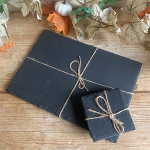 May include: Two black slate serving boards, one rectangular and one square, tied with twine. The boards are displayed on a wooden surface, with decorative autumn foliage and a small white pumpkin in the background.
