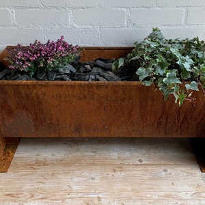 May include: A rectangular, weathered metal planter with a rusty finish. It contains dark grey stones and two plants: a purple flowering plant and a green leafy plant. The planter sits on a wooden surface. The planter's dimensions are approximately 100cm in length.